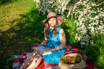 A young lady with a book in her hands on a picnic in the park squints from the sun