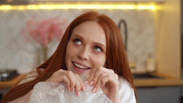 Close-up face of calm young woman playing pops transparent bubble wrap to calm herself sitting at table in kitchen room. Front view of happy smiling redhead female popping plastic bubblewrap.