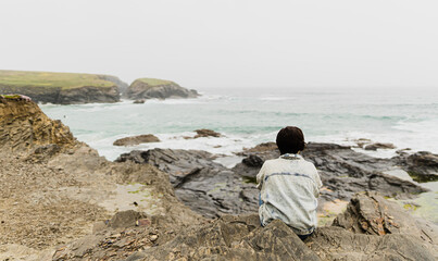 Attractive young female sitting on top of a rock and witnessing the waves hitting the rocks of the cliffs during a cloudy summer day at Cornwall