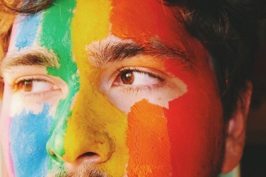 Closeup face portrait of queer young man painted in lgbtq rainbow pride flag 
