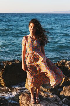 Portrait of woman of turkish ethnicity standing on rocks behind the sea ocean sunset background with floral dress & direct sunlight 