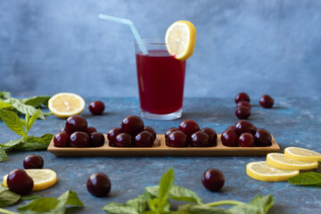 Ripe cherries on wooden board in front of cherry juice glass with straw. Cherries, mint leaves and lemon scattered around. Blue background.