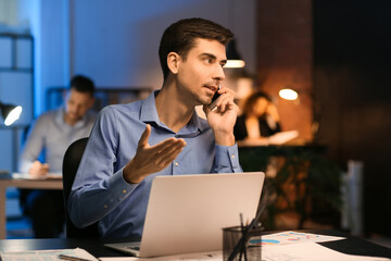 Young man talking by mobile phone in office at night