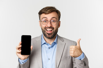 Close-up of excited businessman in gray suit and glasses, showing thumbs-up and mobile phone screen, showing something amazing, standing over white background