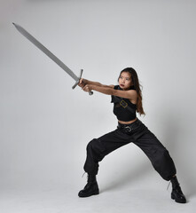 
Full length portrait of pretty young asian girl wearing black tank top, utilitarian  pants and leather boots. Standing pose  holding a sword, isolated against a  studio background.