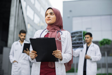 Pretty smiling Arab female doctor in hijab poses outdoors in front of modern clinic and looks away. Two confident male doctors standing behind and working with MRI scan and tablet pc