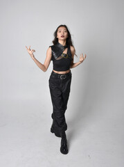 Full length portrait of pretty young asian girl wearing black tank top, utilitarian  pants and leather boots. Standing pose with gestural hands, isolated against a  studio background.