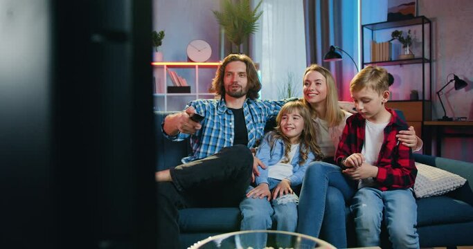 Portrait of adorable happy positive modern family which resting on the couch at home in the evening and having fun together while watching interesting show on TV set