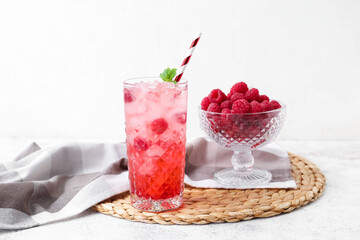 Glass of tasty lemonade and bowl with raspberry on light background