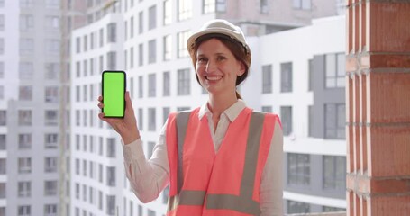 Caucasian woman engineer shows green phone screen, standing in a helmet, smiling. Engineer holds the phone with the app and looks at camera.