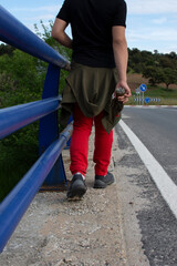 Teenager walking on the shoulder of a road crossing a bridge with blue handrail with a plastic bottle in his hand.