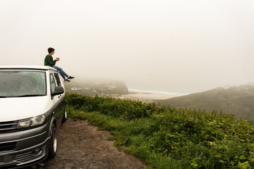 Attractive young female sitting on the roof of a campervan while enjoying the views from the top of...