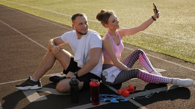 A couple in sportswear takes a selfie while sitting on a treadmill at a stadium.
