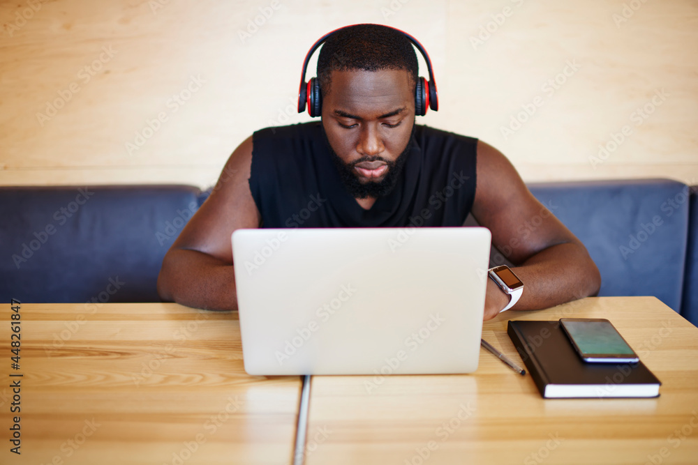 Poster dark skinned male blogger typing program code on digital netbook while listening music playlist in b