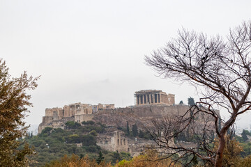 Acropolis hill (Parthenon, Propylaea, Temples, Odeon of Herodes Atticus) with bare tree silhouette. Athens ancient historical landmark in city center from Filopappou Hill on cloudy day