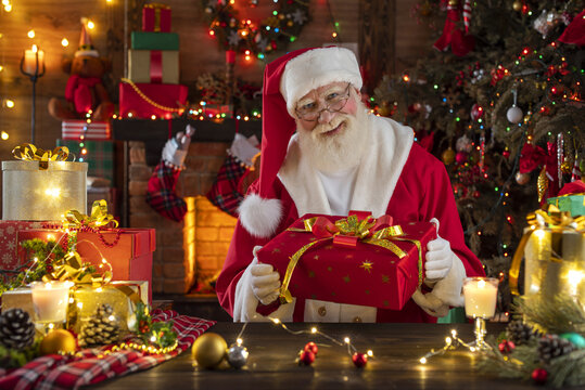 Santa Claus with gift box presents near fireplace and Christmas tree. Festive interior inside wooden house, New Year's cheerful mood Spirit of Christmas. Senior man with real white beard cosplay