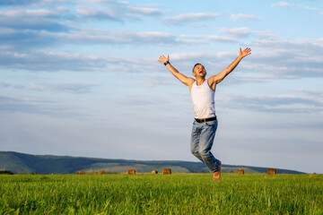 Fototapeta premium A cheerful farmer jumps on a field among hay bales on a hot summer day. People on summer vacation at the farm. Rural landscape in warm colors.