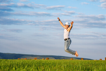 A cheerful farmer jumps on a field among hay bales on a hot summer day. People on summer vacation at the farm. Rural landscape in warm colors.