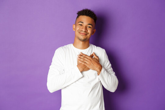 Portrait of happy smiling african-american man, feeling grateful and touched with nice gesture, holding hands on heart, thanking for something, standing over purple background