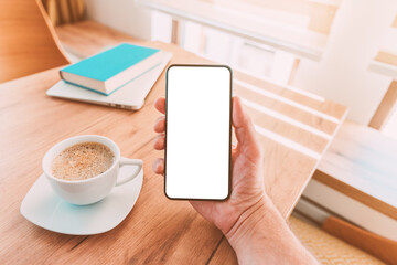 Blank mobile smart phone screen mock up. Man holding smartphone over desk with coffee cup at home.