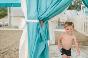 little boy in swimwear in summer stands on the beach under the canopy of a gazebo and smiles