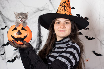 A woman in a witch costume in a hat holds a bucket pumpkin with cat. Halloween people and animal.