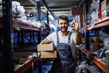 Young smiling happy bearded tattooed hardworking employee holding boxes and standing in storage of import and export firm and showing okay gesture.