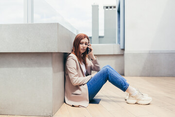 young frustrated woman sitting on sidewalk near a depressed business office center has lost job, financial problems. Female student girl stress at work or university college. Lady with grief outdoors