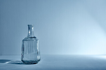 Glass empty open bottle with copy space. Graphic still life with light and shadow in blue light. The concept of glass containers, recyclable materials or alcoholism. Large creative vase. Open flask