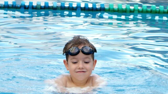A preschool child in the pool in swimming goggles splashes on the water in the pool, a boy jumps and holds a swimming board in his hands.