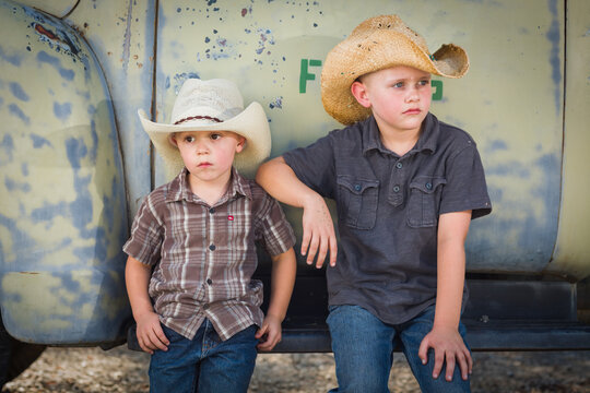 Two Young Boys Wearing Cowboy Hats Leaning Against an Antique Truck in a Rustic Country Setting..