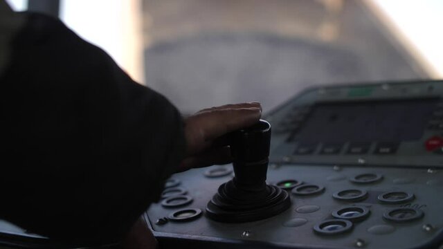 Close-up view of hand of driver or heavy equipment operator sitting at control panel and using joystick inside cabin of road paving machine against hot asphalt falling from dump truck in background.