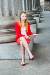 Dressing in a red blazer, white under wear, a red skirt and open toes high heels, crossing legs, a young blonde teenage girl is sitting on a column base, studying on a tablet computer..
