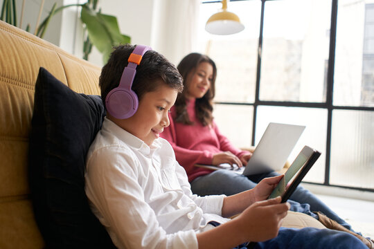 Boy with headphones playing with a tablet on the sofa at home while his mother works remotely on her laptop. Concept of remote work and family reconciliation.