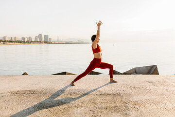 Hispanic mid adult woman doing yoga and meditating by the sea - Fitness woman practicing warrior...