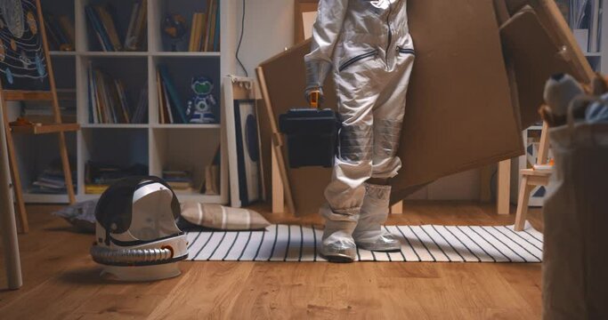 Happy little child wearing astronaut suit enters room with big cardboard piece and tool box to build rocket space ship.