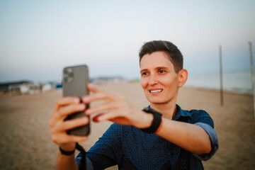 An androgynous woman with short hair smiles and looks at her phone on a beach