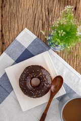 chocolate donut and wooden spoon in white square ceramic plate beside coffee and vase on napkin and rustic natural wood texture background, easy meal for break time, top view