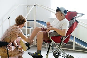 Mother helping disabled boy 17 years old in wheelchair with cerebral palsy to put on shoes, before...