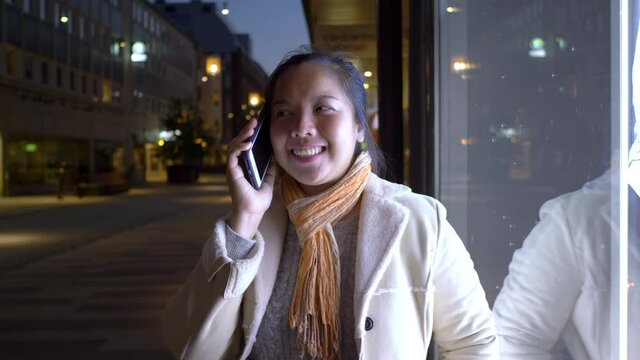 Happy Asian woman talking on mobile phone and tanding against the wall in the city at night. Smiling female using smartphone outdoor. City night concept.