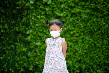 Adorable small girl with healthy face mask standing against green nature background.