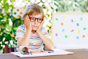 Confused little kid boy with glasses with colorful wax crayon pencils. Sad child and student is...