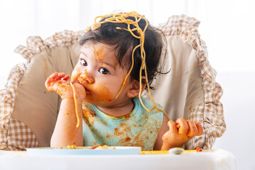 Adorable little child funny girl eating spaghetti with spoon while sitting in high-powered chair at...
