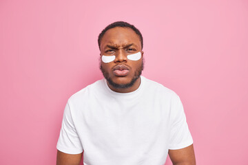 Displeased dark skinned man has sulking expression beard and full lips applies patches under eyes to reduce wrinkles wears basic casual t shirt isolated over pink background. Beauty concept.
