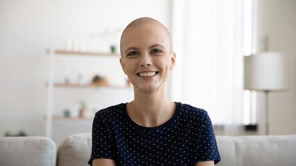 Head shot portrait of happy young bald after chemotherapy woman looking at camera. Smiling sincere millennial hairless lady holding video call with doctor, feeling excited of hearing positive news.