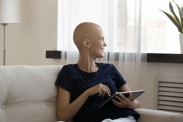 Dreamy positive young hairless woman using digital computer tablet, enjoying playing online games,...