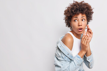 Fototapeta na wymiar Studio shot of impressed curly haired woman looks wondered away stares in complete disbelief wears white t shirt and denim shirt isolated over white background blank copy space for your advert