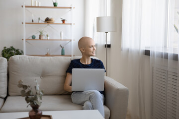 Dreamy young relaxed hairless cancer ill woman sitting on sofa with computer on laps, getting message with good news, looking in distance visualizing healthy future, enjoying carefree weekend time.