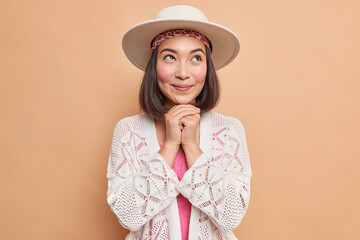 Horizontal shot of dreamy Asian woman keeps hands under chin thinks about something pleasant wears fedora white knitted jumper makes plans in mind isolated over beige background. Pleasant thoughts