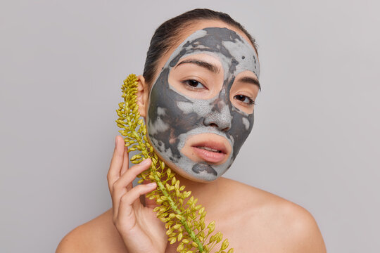 Beauty photo of brunette young Asian woman applies homemade mask on face holds wildflower poses naked indoor isolated over grey studio background. Wellness skin rejuvenation and facial care concept.